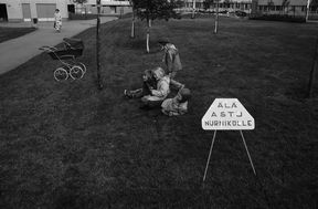 Three children are sitting or lying on the lawn in the yard of an apartment building, while another walks casually across the grass. In the background, a sign reads 'Don't step on the lawn'.