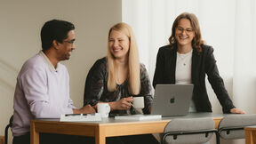 Tre personer diskuterar runt ett bord med en bärbar dator, anteckningsbok och kaffekoppar.