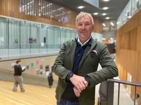 Man in green blazer and dark shirt standing indoors by a railing. Wooden walls and other people present in background.