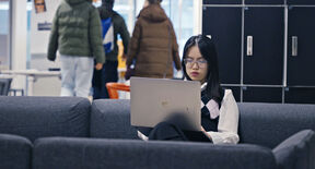 Person sitting on a grey sofa using a laptop in a modern indoor space with people in the background.