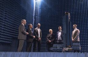 Group of people in a room with blue soundproofing panels, standing around a tall antenna structure.