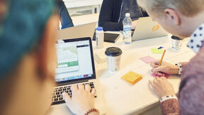 Ihmisiä työskentelemässä pöydän ääressä, kannettavat tietokoneet, muistilaput ja kahvikupit. Yksi henkilö kirjoittaa muistiinpanoja.