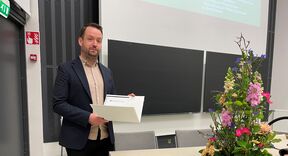 A man holding a certificate stands in front of a chalkboard in a lecture hall. A colourful flower arrangement is on the table.