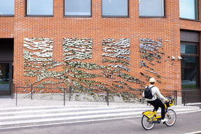 En person cyklar på en gul cykel förbi en tegelvägg med abstrakta metallkonstinstallationer. Cyklisten bär hatt och ryggsäck.