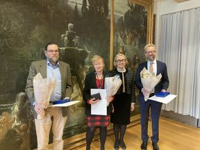 Neljä henkilöä seisoo sisällä kukkia ja todistuksia pitäen. Taustalla koristeellinen maalaus ja vaaleat verhot.