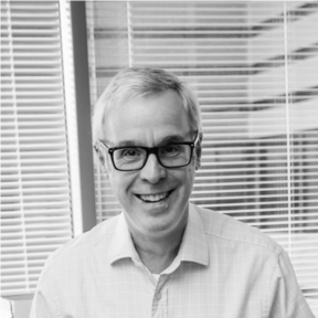 Peter Kelly with grey hair wearing glasses and a light-coloured shirt sits in front of a window with blinds.