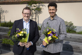 Kaksi miestä, joilla on kukkakimput, seisovat terassilla. Toinen pukeutunut mustaan pukuun ja toinen raidalliseen paitaan.