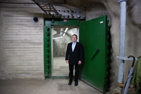 Mies puvussa seisoo suuren vihreän metallisen oven vieressä maanalaisessa bunkkerissa.
