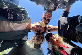 Kuvassa näkyy viisi opiskelijaa aurinkolasit päässä. Opiskelijat seisovat kehässä, ja kuva on otettu sen keskeltä alhaalta päin. 