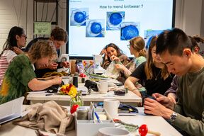 People at a workshop learning to mend knitwear. A presentation screen shows close-ups of knitting repairs.