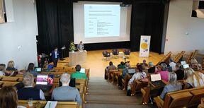 People in a lecture hall are listening to a speaker presenting in front of a screen.