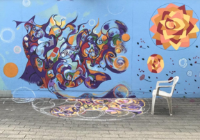 Colorful mural with abstract swirling shapes and geometric patterns on a blue background. In front, a white plastic chair.
