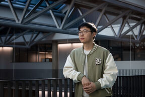 Haoye Tian seisoo käytävällä yllään vaaleanruskea ja valkoinen college-takki, taustalla modernia arkkitehtuuria.