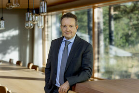 A man in a dark suit and blue tie stands in a wooden room with large windows and hanging lights.