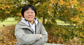 Person standing outdoors in autumn, wearing a grey hoodie and green jacket. Trees in the background with orange leaves.