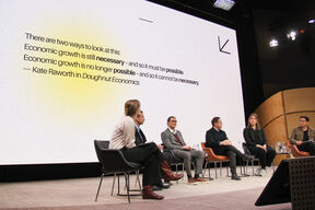 A panel discussion with six people seated on stage. A large screen behind displays a quote about economic growth.