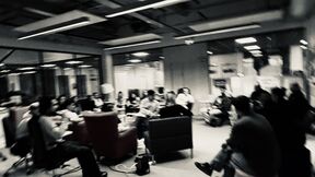 A blurred image of a group of people sitting and discussing in a modern office space with glass walls.