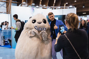 A person poses with a large white furry mascot holding a sign that says 'Sievo' at an event. Another person takes a photo.