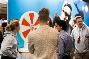 People standing around a red and white spinning wheel at an event. A man in a beige sweater is in the foreground.