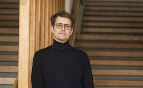 A person in a black turtleneck jumper stands next to wooden railings with stairs in the background.