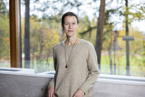 A person wearing a beige sweater and necklace stands indoors by a window with a forest view.