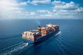 A large cargo ship loaded with colourful containers sails across the blue ocean under a partly cloudy sky.