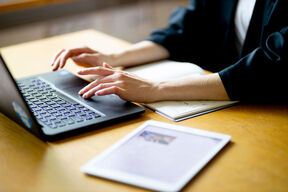 A person typing on a laptop with a notebook and tablet on a wooden desk.