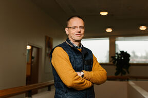A person wearing a yellow sweater and dark blue vest stands with arms crossed in an indoor setting.