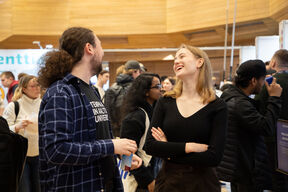 Two people talking at an indoor event. One wears a checked shirt and holds a cup, the other wears a black top with arms crossed.
