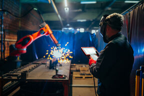 Työntekijä käyttää tablettia, kun robottikäsi hitsaa metallia ja kipinöitä lentää teollisuusympäristössä.