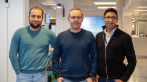 Three men standing indoors, wearing casual clothes. The background includes a screen and office furniture.