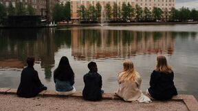 Five people sit by a lake, facing away. Buildings and trees are reflected in the water.