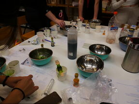 A table with bowls, bottles, and cups. People are working with various items, including a bottle of dark liquid.