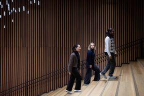 Kolme ihmistä kävelee ylös puisia portaita seinän vieressä, jossa on pystysuorat puusäleet ja roikkuvia koristeita.