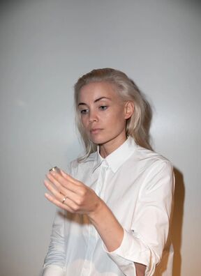 Person with long blonde hair wearing a white shirt, holding a small object in their hand.