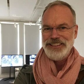 A person wearing a pink scarf indoors with two screens and large windows in the background.