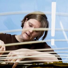 A person weaving thin strips of material. The background is blue, and the person wears a dark top.