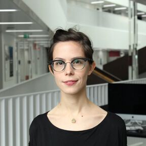 A person with dark hair in a bun wearing a black top and a small pendant necklace in a modern indoor setting.
