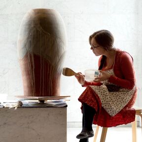 A person in a red dress and apron is painting a large ceramic vase with a brush. The vase is on a table.