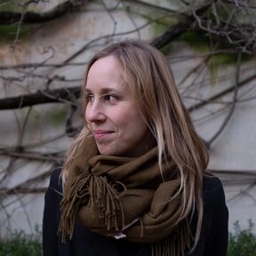 A person with long blonde hair wearing a dark coat and a large brown scarf. The background has tangled branches and a wall.