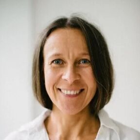 Person with short brown hair wearing a white shirt, standing against a light grey background.