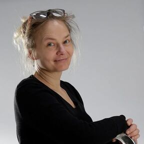 A person with glasses on their head, wearing a black top, arms crossed, against a grey background.