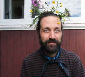 Person wearing a flower crown and a striped shirt with a scarf, standing in front of a red wooden wall and white window.