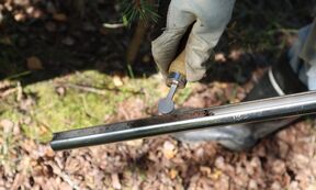 A gloved hand holds a soil sampler with soil inside, over a forest floor covered in leaves and moss.