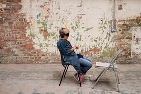 A person wearing headphones sits on a chair facing a folding chair in a room with a worn brick and plaster wall.