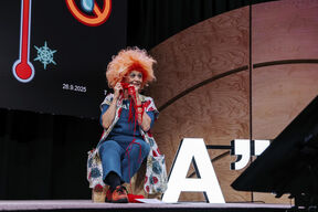 Oranssikiharainen henkilö istuu lavalla pitäen punaista lankaa. Näkyvissä iso näyttö ja puinen tausta.