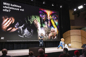 Esitys, jossa suuri näyttö näyttää värikkäitä kuvia ja tekstiä. Kaksi ihmistä lavalla, yksi seisoo ja yksi istuu.