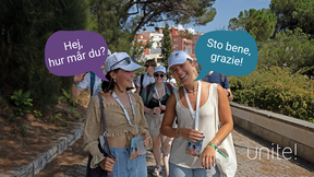 En grupp människor går utomhus, två kvinnor längst fram samtalar. En säger 'Hej, hur mår du?' och den andra svarar 'Sto bene, grazie!'.