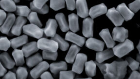 A close-up of numerous small, rectangular particles with rounded edges, appearing grey on a dark background.