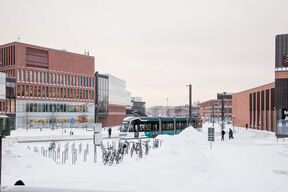 A snowy urban scene with modern buildings, a tram, and bicycles parked. People walk along the snow-covered paths.